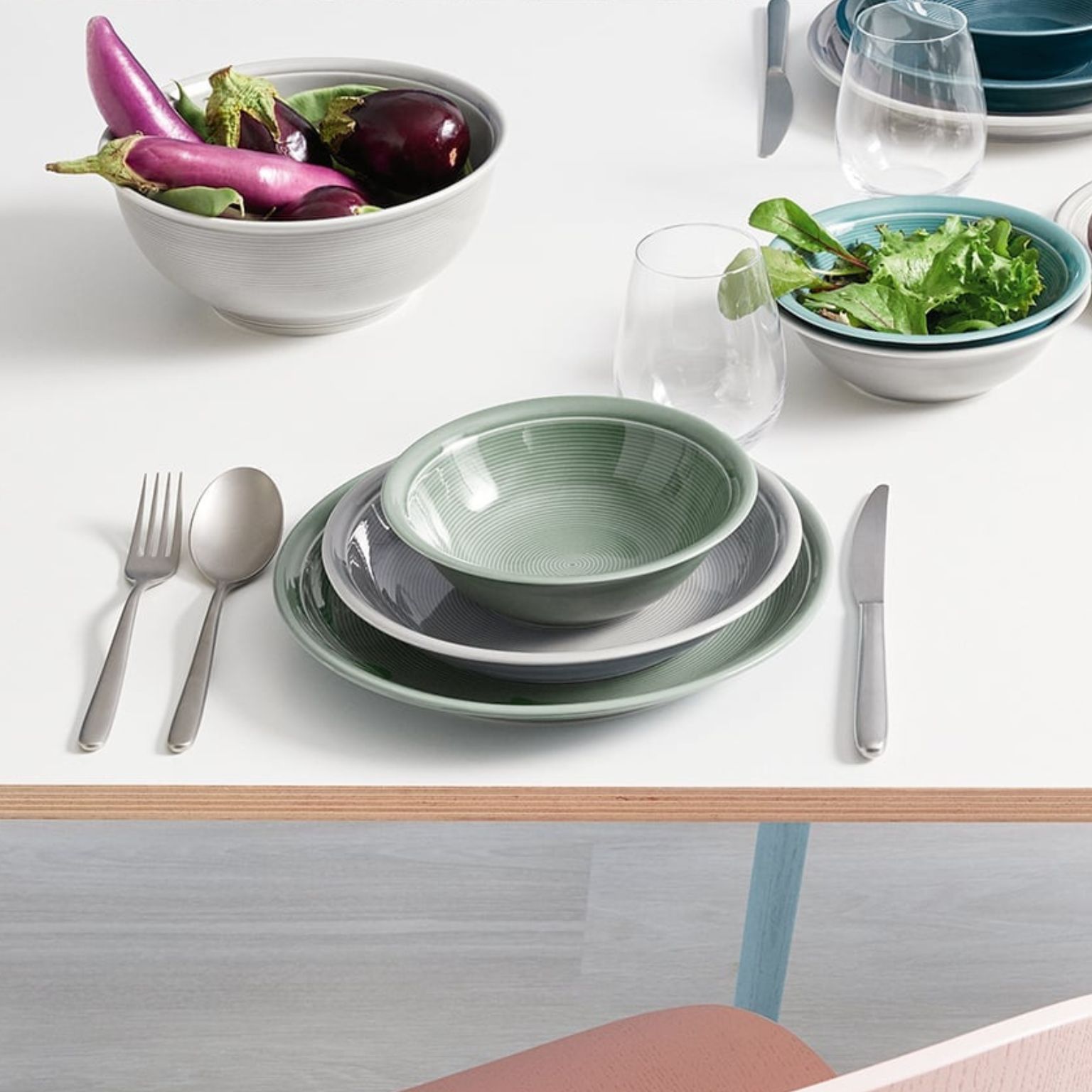 A modern table setting with green-toned dishware, including plates, bowls, and stainless steel cutlery. Fresh salad and eggplants are in bowls, with glasses and additional tableware in the background.